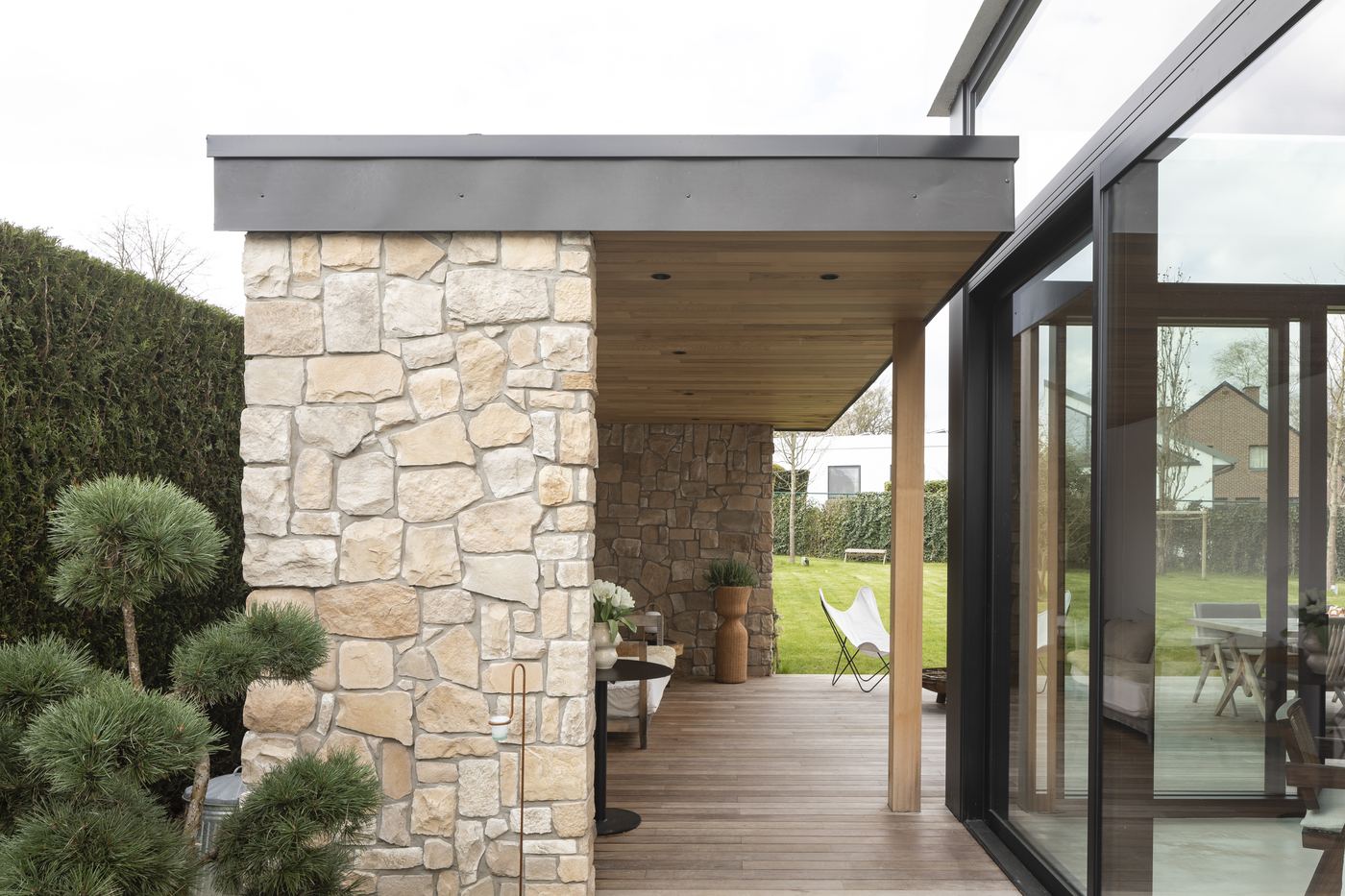 Terrasse couverte avec mur en pierre naturelle, plafond en bois et grandes baies vitrées